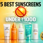 A wide horizontal product showcase banner for a skincare blog, dimensions 1200x628. Split background: left side is a bright sunny yellow-orange gradient, right side is a cool mint green-blue gradient. In the center, five modern sunscreen product bottles lined up in a row, photorealistic style, looking premium and glossy. Above the products, a large glowing sun icon with a red "prohibited" (circle with slash) symbol over it floating in the air. Bold typography in the foreground: Main text "5 BEST SUNSCREENS" in huge black bold font at the top center. Below it, in a large stencil style, "UNDER ₹1000" in white with a slight shadow. At the bottom right, a circular gold badge that says "2026's Top Picks". A subtle arrow graphic pointing from the text towards the products. High contrast, vibrant colors, magazine quality, 4k, sharp focus, commercial product photography style.